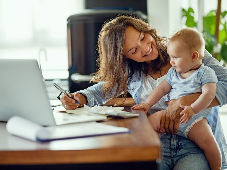 Eine Frau im Homeoffice mit ihrem Baby auf dem Arm sitzt vor ihrem Laptop.