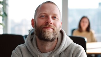 Ein Mann mittleren Alters mit Bart und Hoodie schaut direkt in die Kamera. Im Hintergrund sitzt eine Frau an einem Tisch