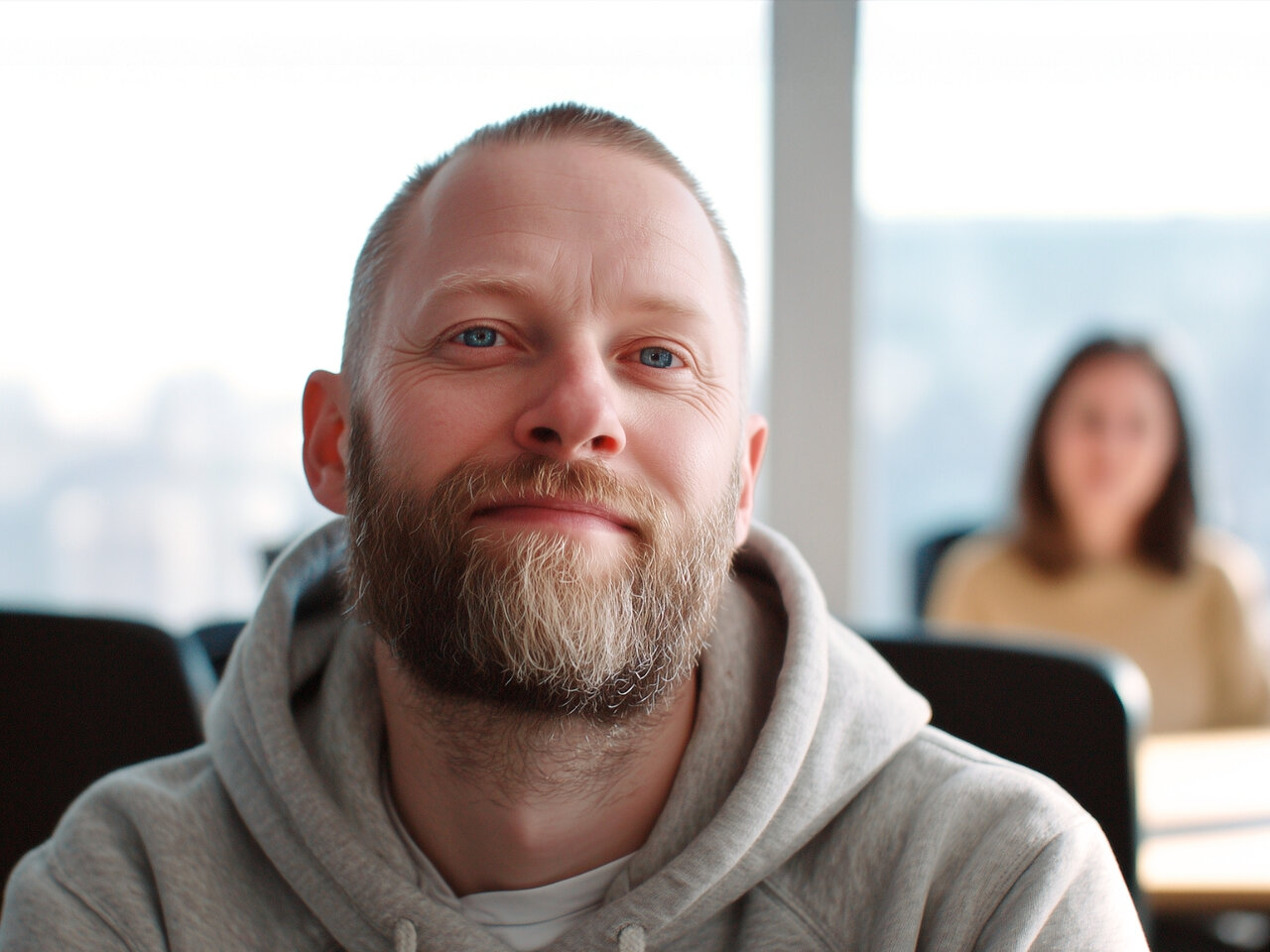 Ein Mann mittleren Alters mit Bart und Hoodie schaut direkt in die Kamera. Im Hintergrund sitzt eine Frau an einem Tisch