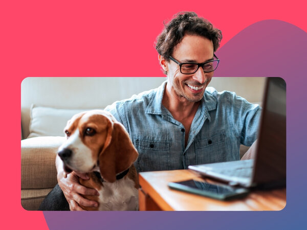 Mann mit Brille sitzt lächelnd auf dem Fußboden, an ein Sofa angelehnt, vor ihm ein Laptop auf einem Couchtisch, ein Hund sitzt neben ihm.