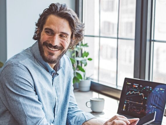 Mann mit Diagrammen und Kursen Ein Finanzexperte schaut sich Diagramme und Kurse auf seinem Laptop an und tippt in sein Smartphone