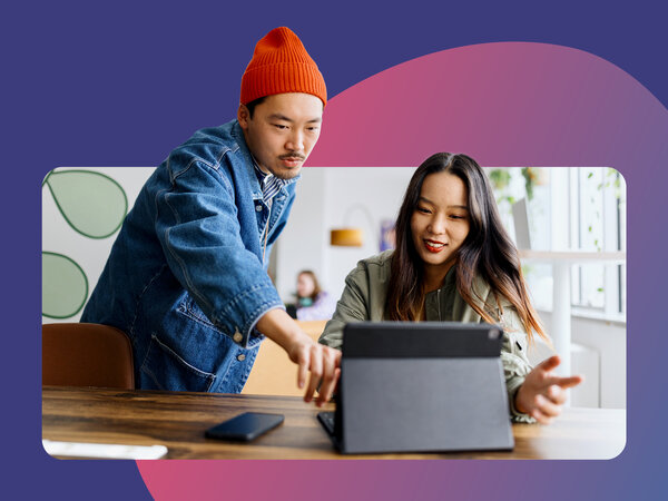 Eine Frau sitzt am Tisch vor einem Laptop, ein Mann steht neben ihr und erklärt ihr etwas.