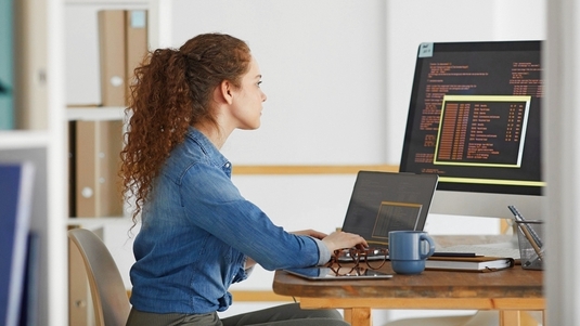 Eine Frau sitzt an einem Schreibtisch mit Laptop und Monitor und arbeitet an Programmiercodes