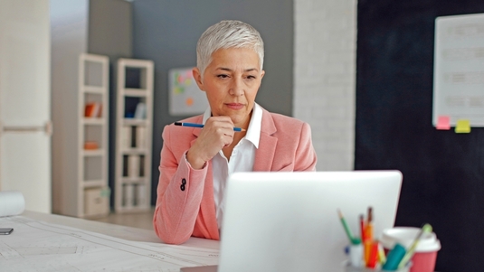 Eine Frau sitzt am Schreibtisch vor Konstruktionszeichnungen und ihren Laptop.