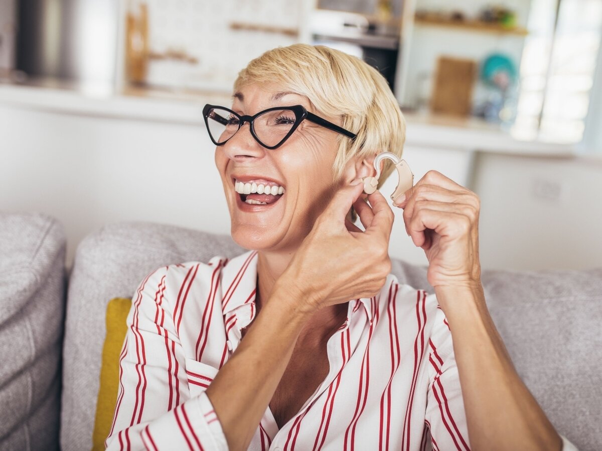 Eine blonde Frau mit Brille, die ihr Hörgerät anzieht und fröhlich lacht