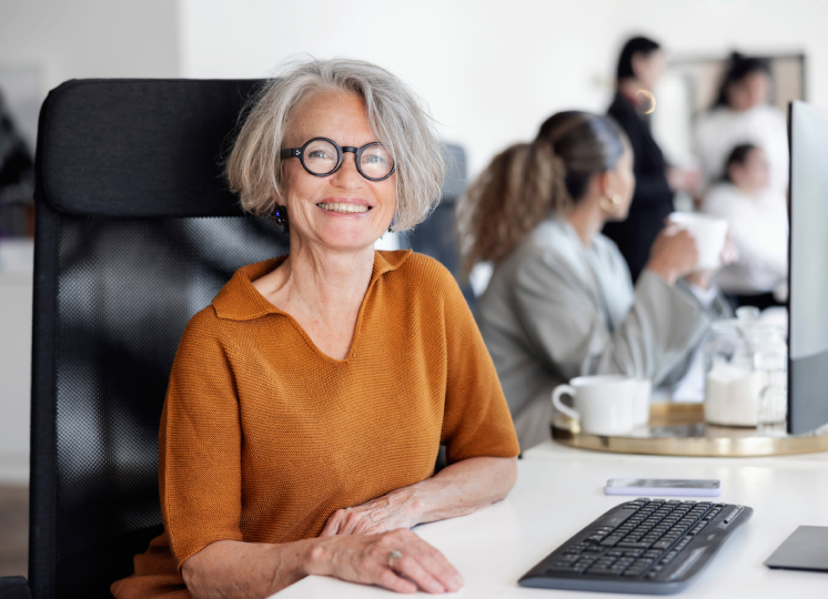Frau mit grauen Haaren und moderner Brille sitzt in einem Buero vor ihrem Laptop und schaut froehlich in die Kamera.