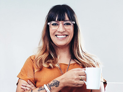 Eine Frau mit Brille und Kaffeetasse in der Hand lächelt freundlich in die Kamera